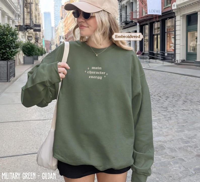A woman wearing a green sweatshirt with embroidered text, sunglasses, and a cap is standing on a city street.