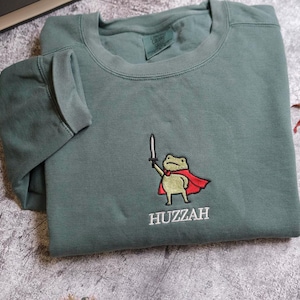 May include: A sage green sweatshirt featuring an embroidered cartoon frog holding a sword and wearing a red cape. The word "Huzzah" is embroidered below the frog. The sweatshirt is folded on a textured surface.