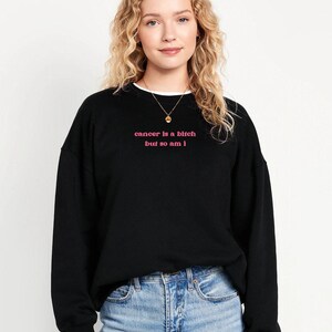 A young woman with curly blonde hair wearing a black sweatshirt with the text "cancer is a bitch but so am I" printed on it.