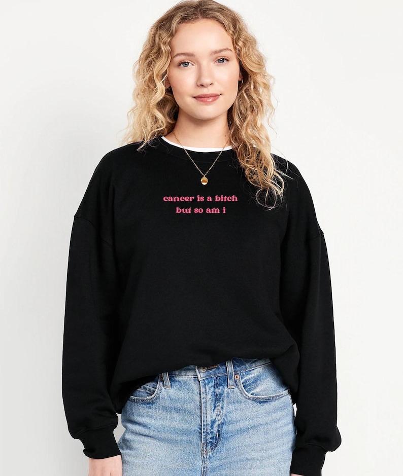 A young woman with curly blonde hair wearing a black sweatshirt with the text "cancer is a bitch but so am I" printed on it.