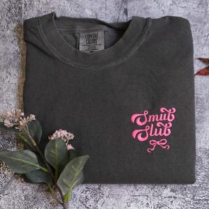 a gray t-shirt with pink text and a small leaf on a textured surface.