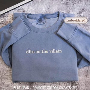 A blue sweatshirt with the text "dibs on the villain" printed on it, along with a label indicating it is embroidered. The sweatshirt is laid out on a textured surface, and there is a small leaf visible in the background.