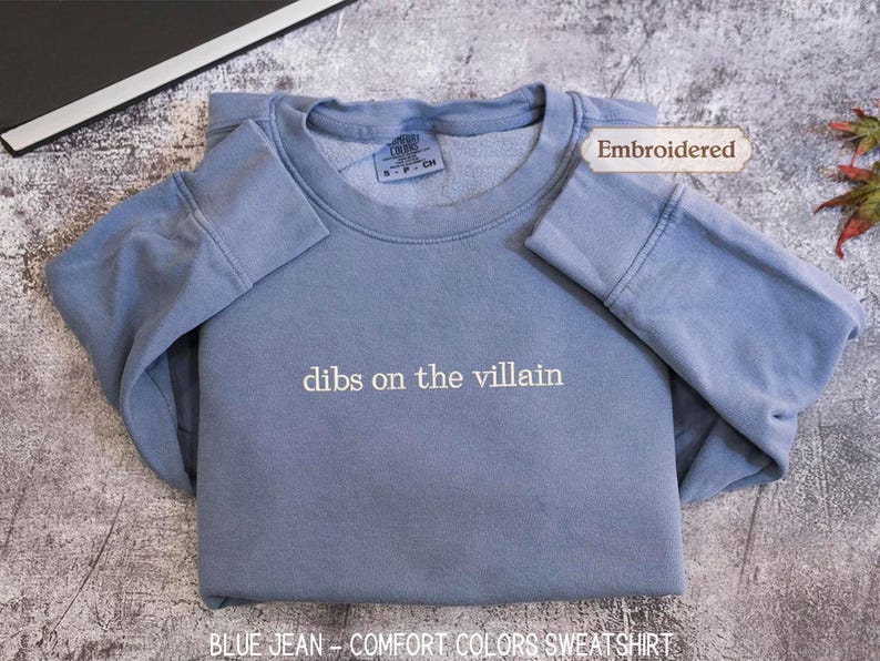 A blue sweatshirt with the text "dibs on the villain" printed on it, along with a label indicating it is embroidered. The sweatshirt is laid out on a textured surface, and there is a small leaf visible in the background.