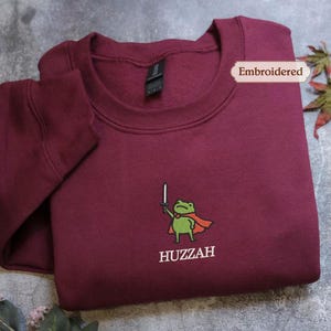 A burgundy sweatshirt with a frog character and the word "HUZZAH" printed on it, along with a small embroidered label.