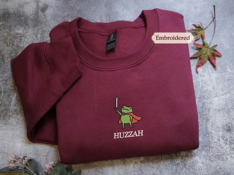 A burgundy sweatshirt with a frog character and the word "HUZZAH" printed on it, along with a small embroidered label.