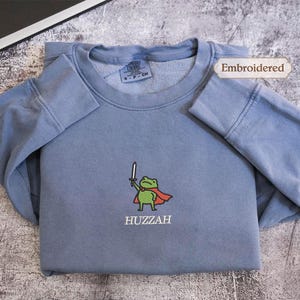 A gray sweatshirt with a frog character and the word "HUZZAH" printed on it, along with a label indicating that it is embroidered.