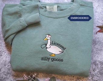 May include: Sage green sweatshirt with an embroidered design of a goose wearing a nurse's hat and stethoscope. The text "Licensed silly goose" is below the goose. The word "EMBROIDERED" is on a navy blue tag.