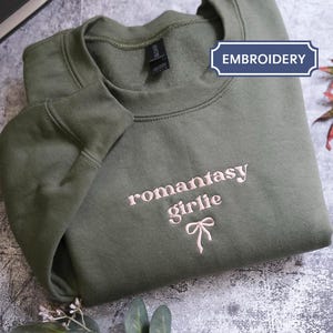 a green sweatshirt with the text "romantic girl" embroidered on it, along with a bow detail. The sweatshirt is folded and placed on a gray surface, with some leaves and a small flower nearby.