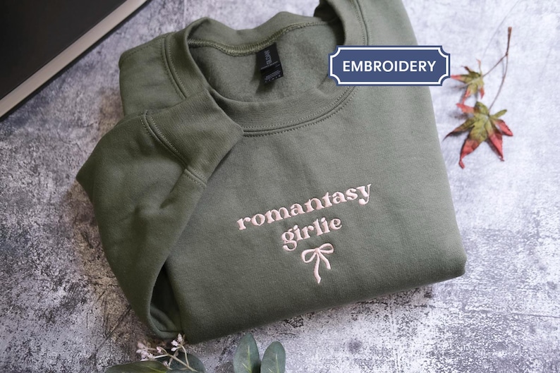 a green sweatshirt with the text "romantic girl" embroidered on it, along with a bow detail. The sweatshirt is folded and placed on a gray surface, with some leaves and a small flower nearby.