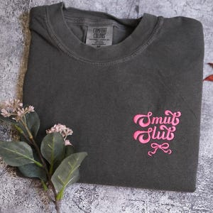 a gray t-shirt with a pink embroidered design, placed on a gray surface alongside some flowers and leaves.