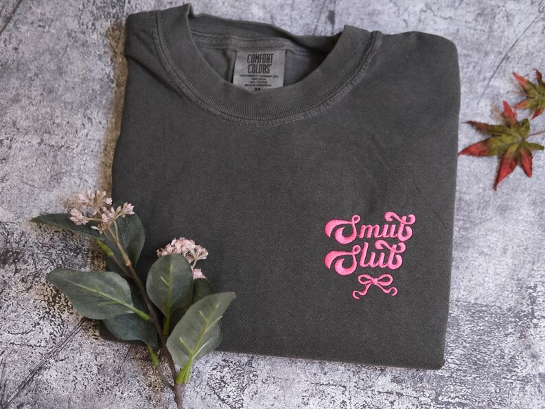 a gray t-shirt with a pink embroidered design, placed on a gray surface alongside some flowers and leaves.