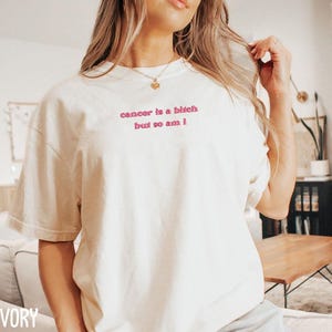 A woman wearing a white t-shirt with the text "cancer is a bitch but so am" printed on it.