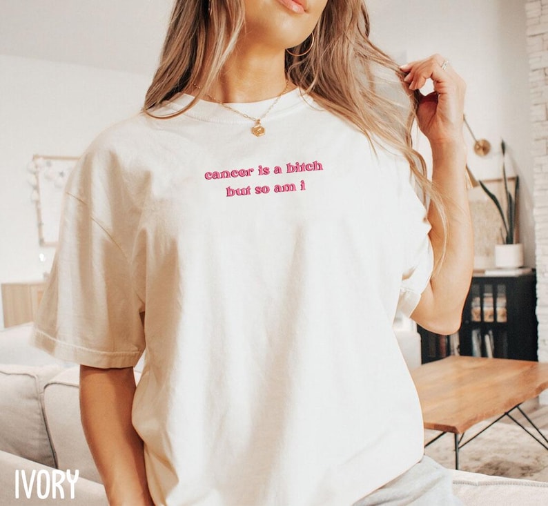 A woman wearing a white t-shirt with the text "cancer is a bitch but so am" printed on it.