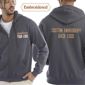 May include: Dark grey zip-up hoodie with a front pocket and a hood. The front of the hoodie has the text "custom YOUR LOGO" embroidered in a gold colour. The back of the hoodie has the text "CUSTOM EMBROIDERY BACK LOGO".