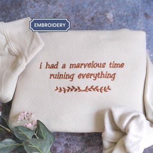 May include: Cream-colored hoodie with the embroidered phrase "i had a marvelous time ruining everything" in a copper-colored thread. A decorative leaf design is embroidered below the text. The hood is visible on the left side.