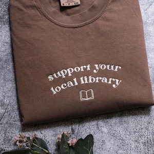 A brown t-shirt with the text "Support your local library" printed on it, along with an open book icon, is displayed on a gray surface.