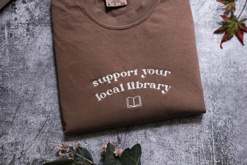 A brown t-shirt with the text "Support your local library" printed on it, along with an open book icon, is displayed on a gray surface.