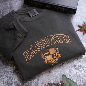 a grey sweatshirt with the word "BASGLATH" printed on it, along with a black book titled "FOURTH WING" and a leaf on the ground.