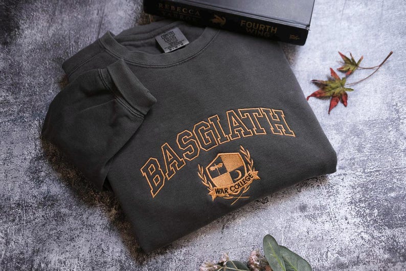 a grey sweatshirt with the word "BASGLATH" printed on it, along with a black book titled "FOURTH WING" and a leaf on the ground.