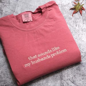 May include: A dusty rose Comfort Colors t-shirt with the phrase "that sounds like my husbands problem" embroidered in white. The shirt is folded and lies on a textured surface, with a disco ball and foliage in the background.