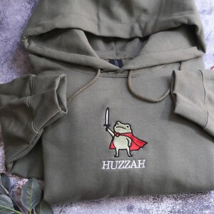 May include: Olive green hooded sweatshirt featuring an embroidered frog wearing a red cape and holding a sword. The word "Huzzah" is embroidered below the frog. The sweatshirt has a drawstring hood and ribbed cuffs.