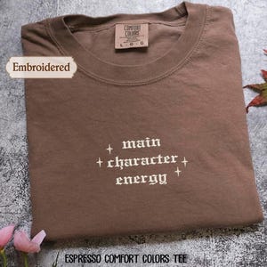 a brown t-shirt with white text printed on it, placed on a gray surface. There are some pink flowers and a leaf in the background.