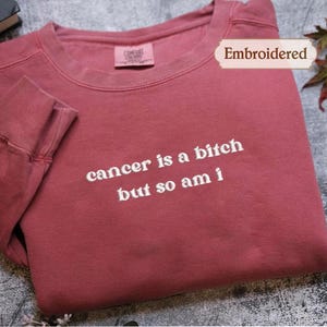A red long-sleeved shirt with the text "cancer is a shit but so am i" printed on it, along with a label that says "Embroidered".