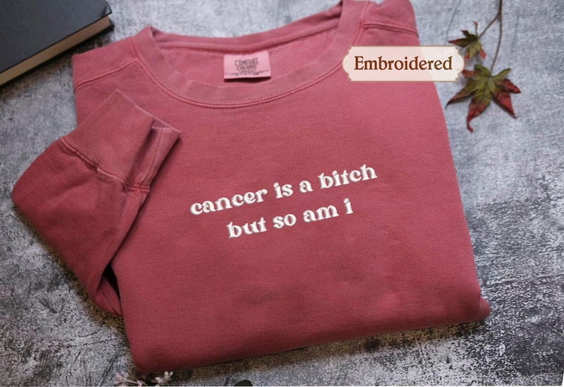 A red long-sleeved shirt with the text "cancer is a shit but so am i" printed on it, along with a label that says "Embroidered".