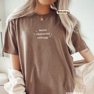 A woman wearing a brown t-shirt with the text "main character energy" printed on it.
