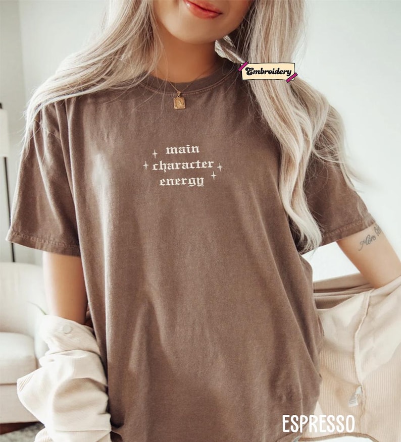 A woman wearing a brown t-shirt with the text "main character energy" printed on it.