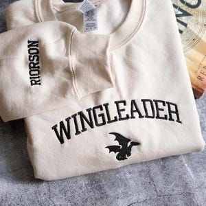 May include: Cream-coloured sweatshirt featuring the word "WINGLEADER" in black arched lettering and a black dragon graphic. The sleeve reads "RIORSON" in black. A book with the title "REBECCA YARROS" is visible.