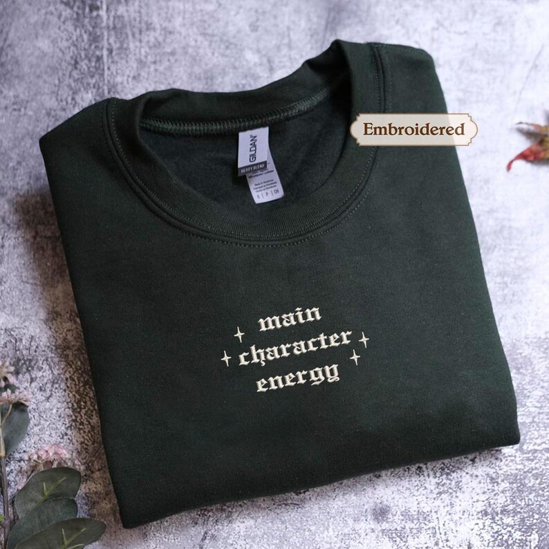 A dark green sweatshirt with the text "main character energy" embroidered on it.