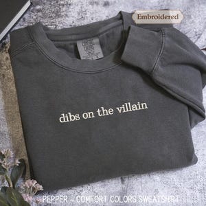 A gray sweatshirt with the text "dibs on the villain" printed on it, along with a label indicating it is "Embroidered" and "Comfortable Colors Sweatshirt".