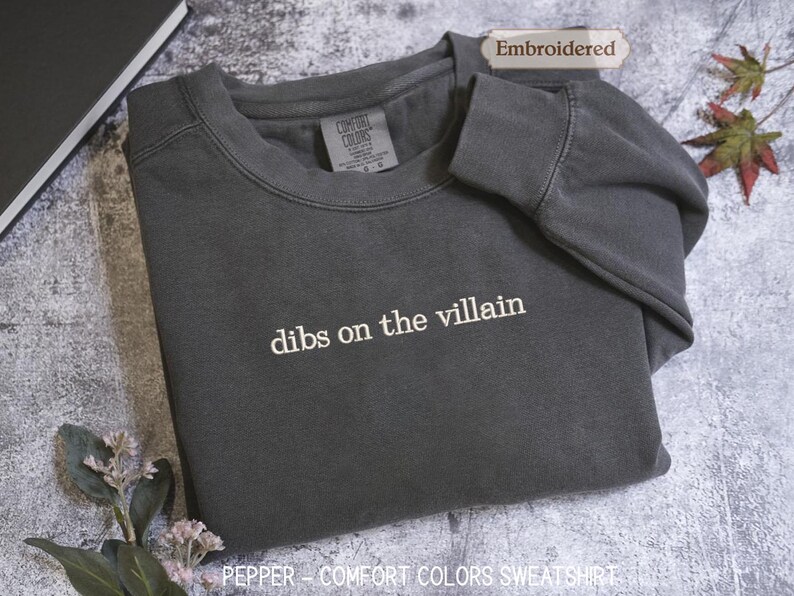 A gray sweatshirt with the text "dibs on the villain" printed on it, along with a label indicating it is "Embroidered" and "Comfortable Colors Sweatshirt".