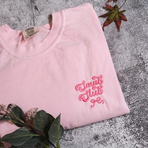 a pink t-shirt with a pink embroidered logo, placed on a gray surface alongside some leaves and a small red leaf.