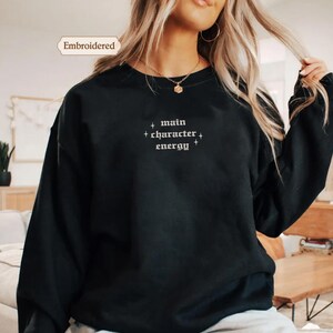A woman wearing a black sweater with the text "main character energy" embroidered on it.
