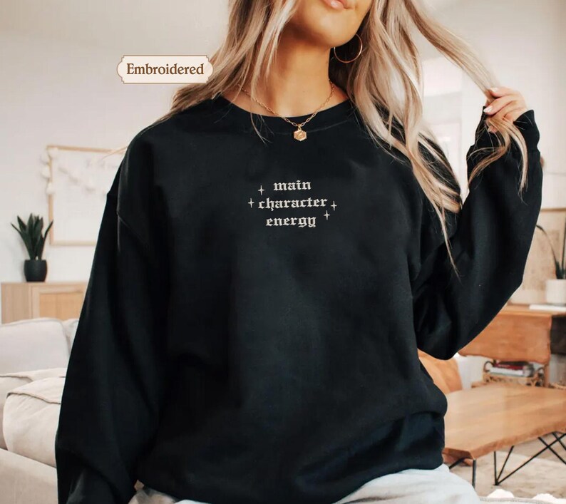 A woman wearing a black sweater with the text "main character energy" embroidered on it.