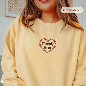 A woman wearing a yellow sweater with a heart-shaped embroidered design and the text "Morally Grey" printed on it.