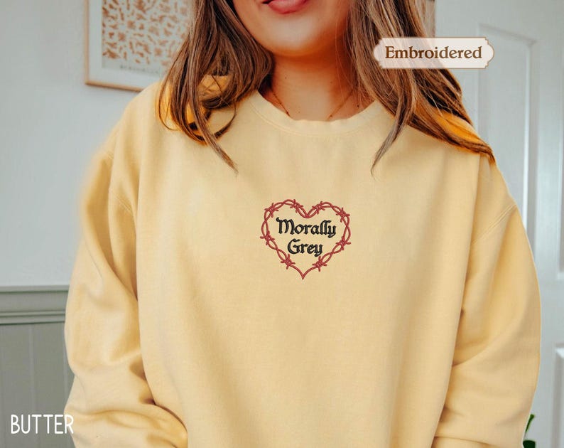 A woman wearing a yellow sweater with a heart-shaped embroidered design and the text "Morally Grey" printed on it.