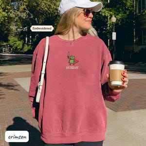 A woman wearing a red sweatshirt with a frog graphic and holding a coffee cup is standing on a brick sidewalk.