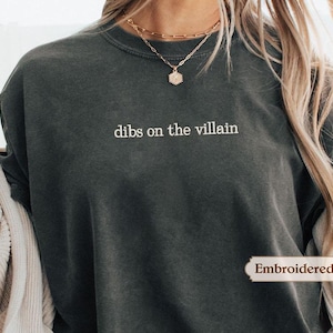 A person wearing a gray t-shirt with the text "dibs on the villain" printed on it, along with a necklace and a sweater.