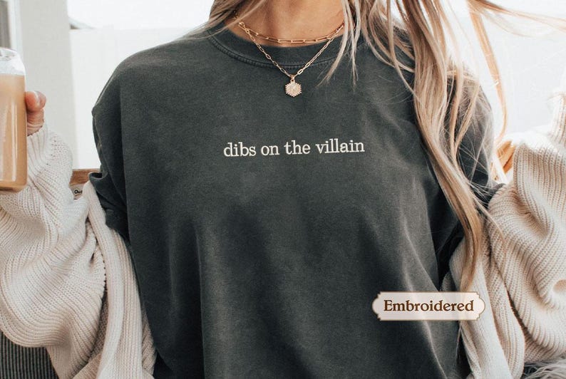 A person wearing a gray t-shirt with the text "dibs on the villain" printed on it, along with a necklace and a sweater.