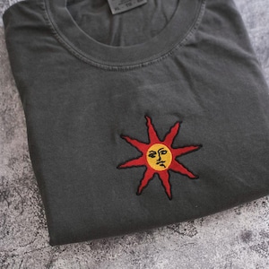 a gray t-shirt with a red sun embroidered on it, resting on a textured gray surface.