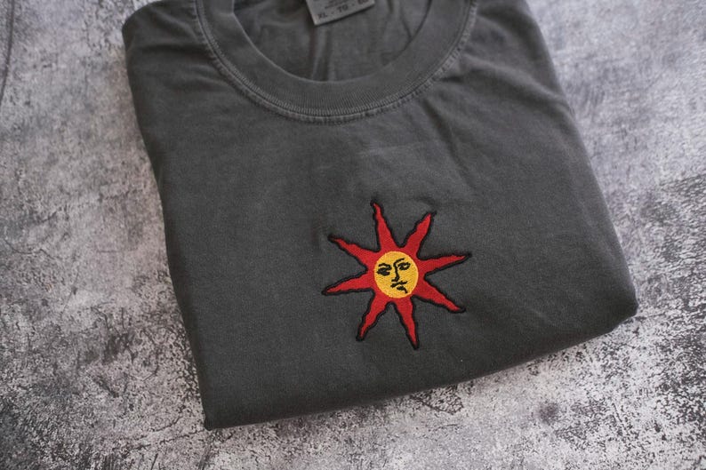 a gray t-shirt with a red sun embroidered on it, resting on a textured gray surface.