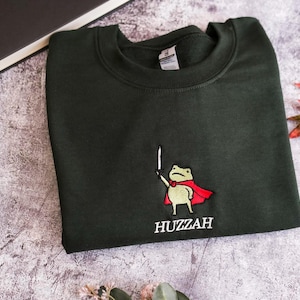 May include: Dark green sweatshirt with an embroidered frog wearing a red cape and holding a sword. The word "Huzzah" is embroidered below the frog. The sweatshirt is folded on a textured surface.