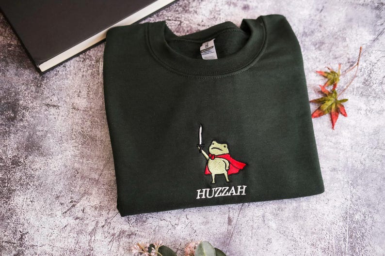 May include: Dark green sweatshirt with an embroidered frog wearing a red cape and holding a sword. The word "Huzzah" is embroidered below the frog. The sweatshirt is folded on a textured surface.