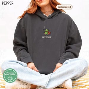 A person wearing a grey hoodie with a frog character and the word "HUZZAH" printed on it.
