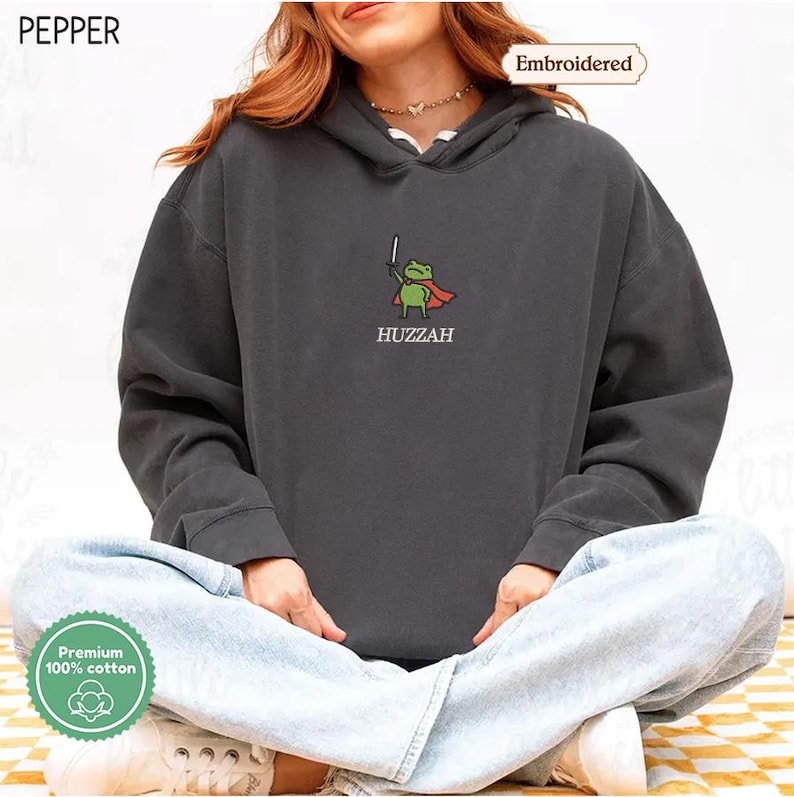 A person wearing a grey hoodie with a frog character and the word "HUZZAH" printed on it.