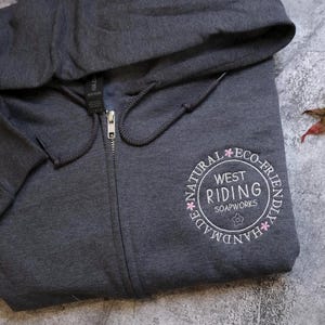 A gray hooded sweatshirt with a circular logo on the front, placed on a gray surface next to a fallen leaf.