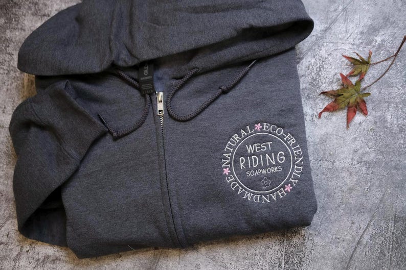 A gray hooded sweatshirt with a circular logo on the front, placed on a gray surface next to a fallen leaf.
